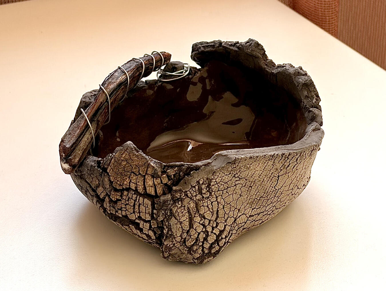 A handcrafted ceramic bowl with natural wood and wire detail, inspired by organic textures and earthy forms. The cracked, bark‑like exterior contrasts with the glossy dark interior, creating a sculptural piece that blends ceramics, nature and contemporary